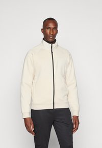 Giacca zip-up beige chiaro con colletto alto, polsini elastici e tasca frontale; realizzata in tessuto morbido con una texture liscia.