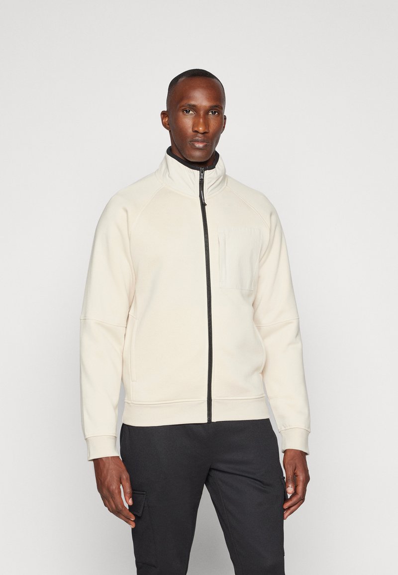 Giacca zip-up beige chiaro con colletto alto, polsini elastici e tasca frontale; realizzata in tessuto morbido con una texture liscia.