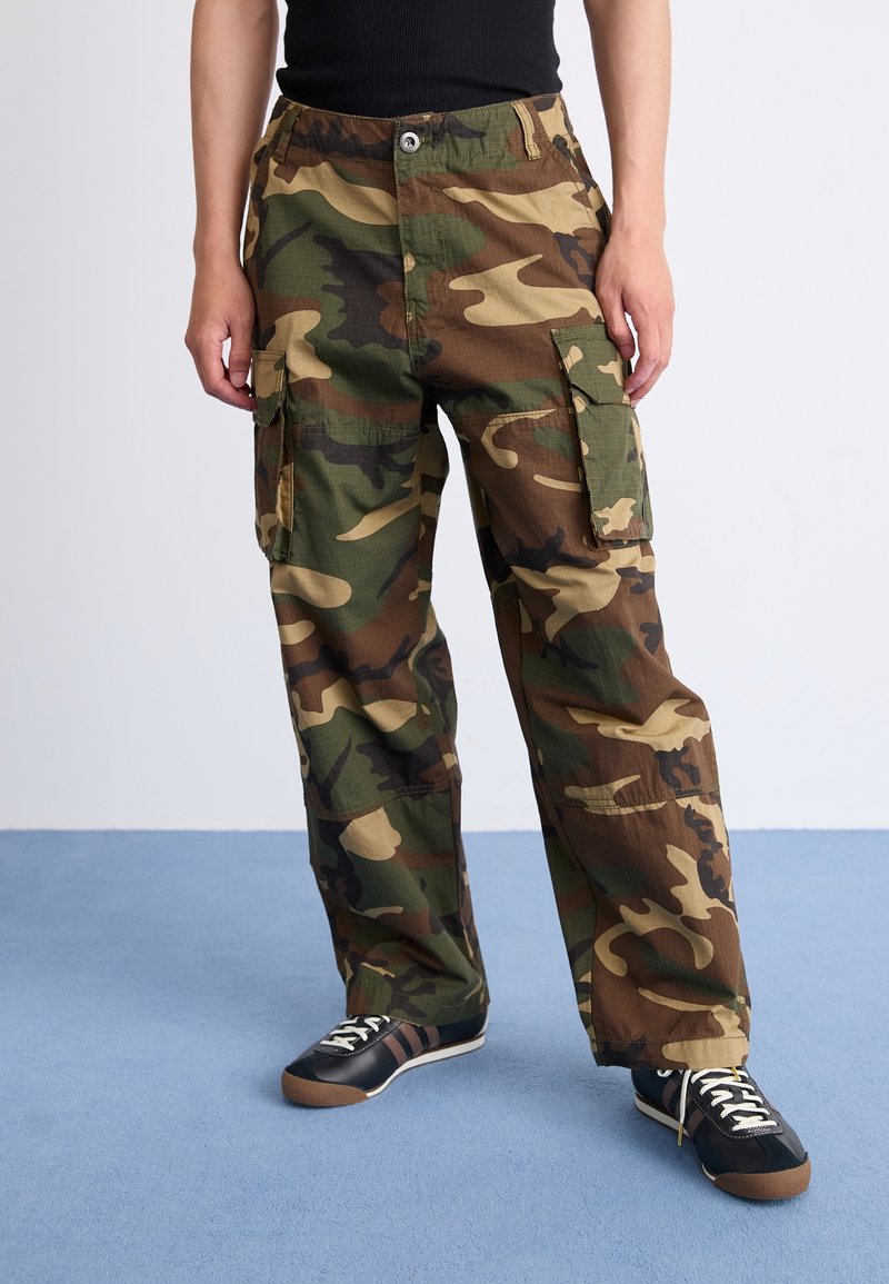 Personne portant un pantalon cargo camouflage vert et marron avec de multiples poches, des baskets noires et un haut noir sans manches.