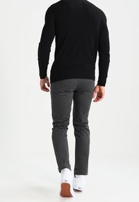 Grijze, strakke broek met een gladde textuur, opgerolde zoom en witte sokken, gecombineerd met een zwart shirt met lange mouwen, gezien van achteren.