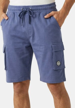 Homme portant un short cargo bleu avec taille à cordon et poches latérales à rabat, les mains dans les poches, sur un fond uni.