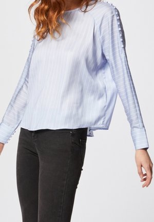 Blusa azzurra chiara a maniche lunghe con strisce bianche verticali, caratterizzata da un collo rotondo e dettagli con bottoni sulle maniche. Abbinata a jeans scuri.