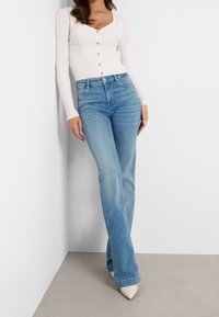 Biała, prążkowana bluzka z długim rękawem, z dekoltem w serek i guzikami, połączona z jasnoniebieskimi, wysoko osadzonymi dżinsami w stylu dzwonów oraz białymi, szpiczastymi szpilkami.