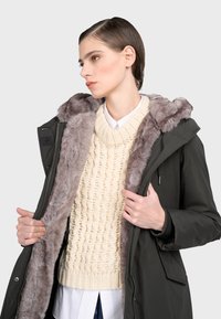 Zielona parka z kapturem wyłożonym futrem i podszewką, noszona na kremowym swetrze w warkocze. Model trzyma parkę otwartą, ukazując teksturę i kontrast kolorów.