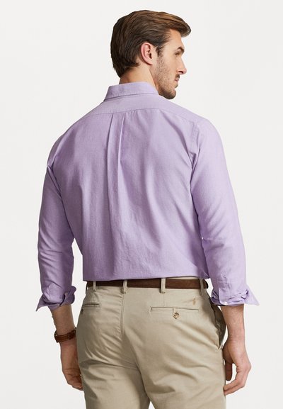 Λεβάντα πουκάμισο με κουμπιά και μακριά μανίκια, με κλασικό γιακά και τυλιγμένους καρπούς, συνδυασμένο με ανοιχτόχρωμο παντελόνι και καφέ ζώνη.