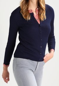 Cárdigan de punto azul marino con un patrón texturizado, escote en v y botones al frente. Se lleva sobre una camisa rosa con cuello y se combina con vaqueros gris claro.