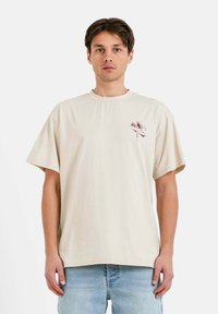 Camiseta de algodón beige con cuello redondo, mangas cortas y un diseño gráfico floral en el lado izquierdo del pecho. Llevada con jeans de color azul claro.