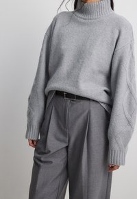 Pull gris clair surdimensionné avec un col roulé côtelé, orné de détails en torsades sur les manches. Associé à un pantalon gris taille haute.