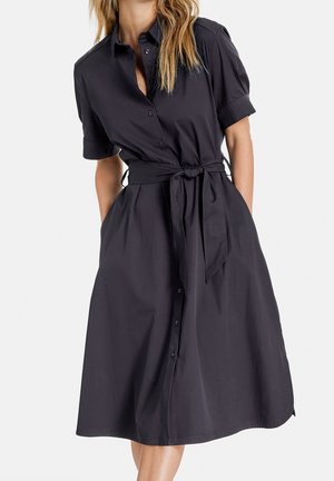 Kvinde iført en sort, kortærmet, knap-ned kjole med et bundet bælte i taljen og lommer, stående foran en ensfarvet baggrund.