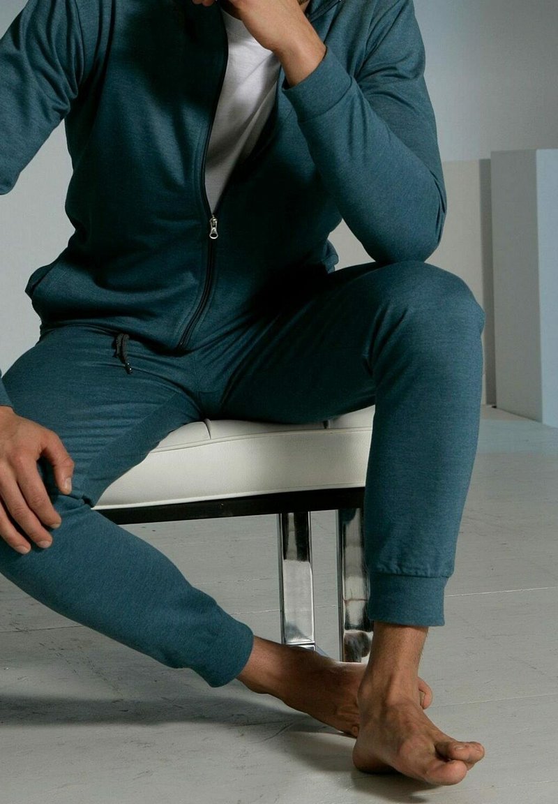 Sudadera con cremallera en verde oscuro y pantalones de chándal a juego, hechos de una tela suave, con cinturilla ajustable y puños acanalados, sentados en una silla blanca.