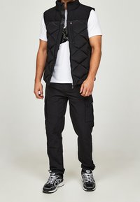 Černá prošívaná vesta s diamantovým vzorem, zapínáním na zip a vysokým límcem, nošená přes bílé tričko, v kombinaci s černými nákladními kalhotami a teniskami.