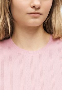 Νεαρή γυναίκα με ξανθά μαλλιά που φοράει ένα ανοιχτό ροζ πλεκτό τοπ με υφή, εμφανίζοντας τον λαιμό και το κάτω μέρος του προσώπου της πάνω σε ουδέτερο φόντο.