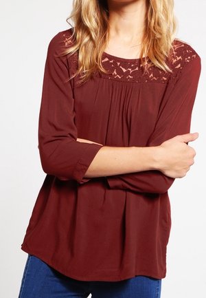 Femme portant un chemisier bordeaux à manches longues avec un détail en dentelle au niveau du col, bras croisés, et un jean bleu visible en dessous.