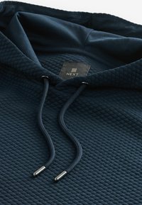 Gros plan sur un sweat à capuche matelassé bleu marine avec un cordon et une étiquette noire indiquant Next à l'intérieur du col.