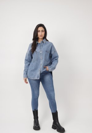 Kvinde stående med den ene hånd i lommen, iført en oversized denimjakke med pletter, tætsiddende blå jeans og sorte chunky støvler mod en ensfarvet baggrund.
