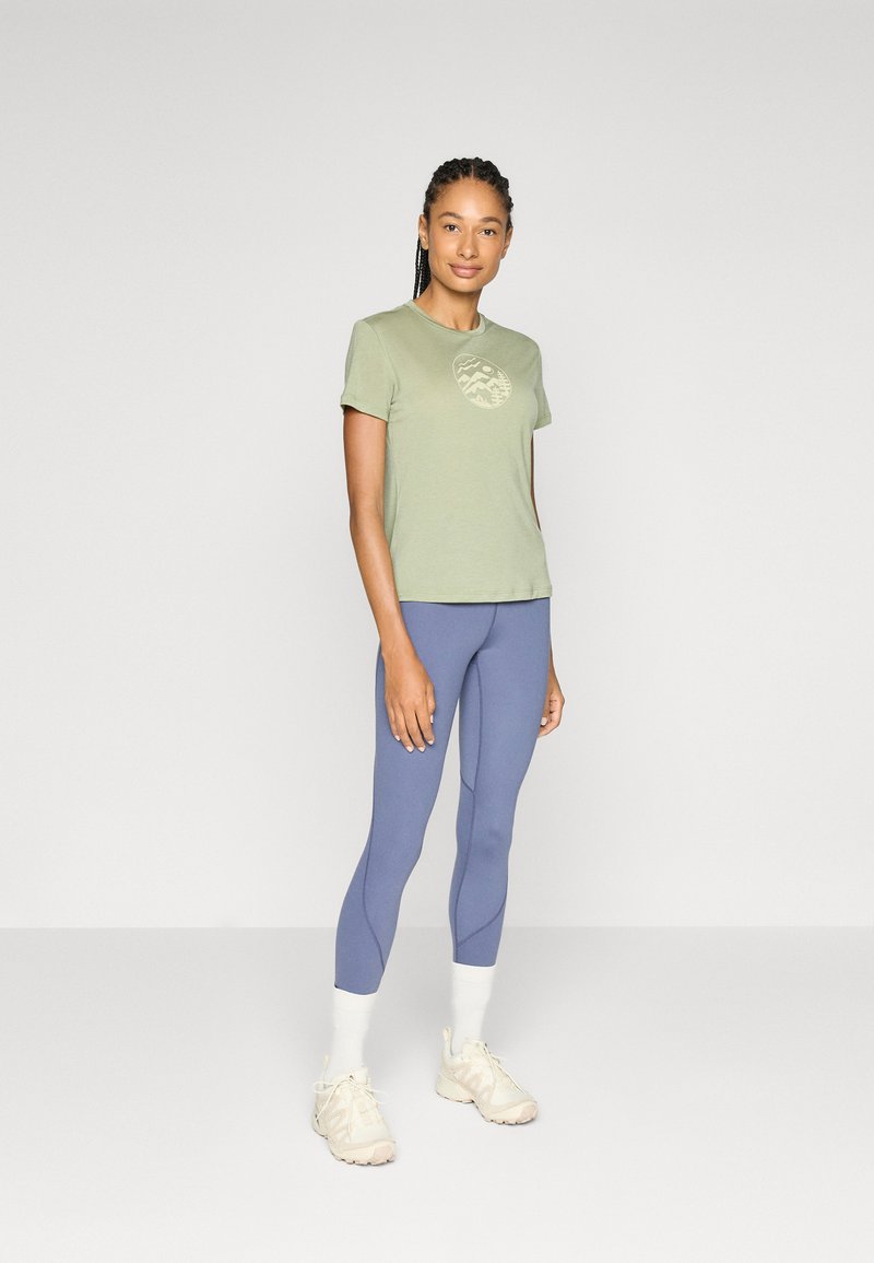 Frau steht in einem hellgrünen grafischen T-Shirt, lila Leggings, weißen Socken und hellbeigen Sneakers vor einem schlichten Hintergrund.