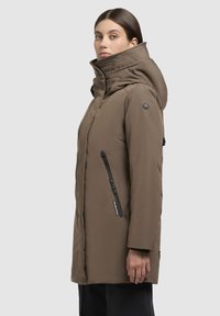 Veste parka rembourrée marron avec un col montant, poches latérales zippées et détail de logo sur l'épaule. Tissu lisse et résistant à l'eau.
