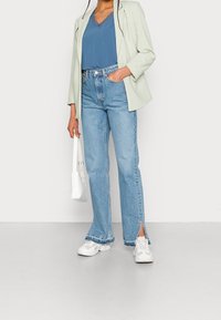 Blazer verde claro, blusa azul con borde de encaje, jeans claros de tiro alto con aberturas laterales y zapatillas blancas, sosteniendo un pequeño bolso blanco.