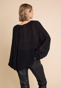 Svart, gjennomskinnelig bluse med rynket hals og brede, luftige ermer. Kombinert med svarte, skinnaktige bukser.
