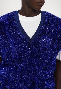Mėlyna be rankovių palaidinė, pagaminta iš tekstūruoto, blizgaus audinio su gilia V formos iškirpte. Po ja vilkima balta marška. Artimas dizaino planas.