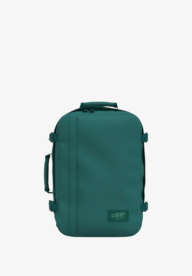 Zaino in tessuto teal di forma rettangolare, con manico superiore, chiusura con zip e patch logo. Texture liscia con cinturini laterali.