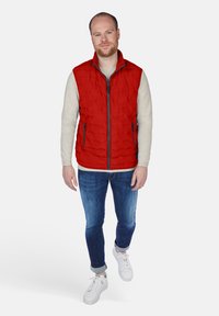 Rote steppte Weste mit vollem Reißverschluss, Seitentaschen und Stehkragen, getragen über einem hellen Pullover und kombiniert mit blauen Jeans und weißen Sneakers.