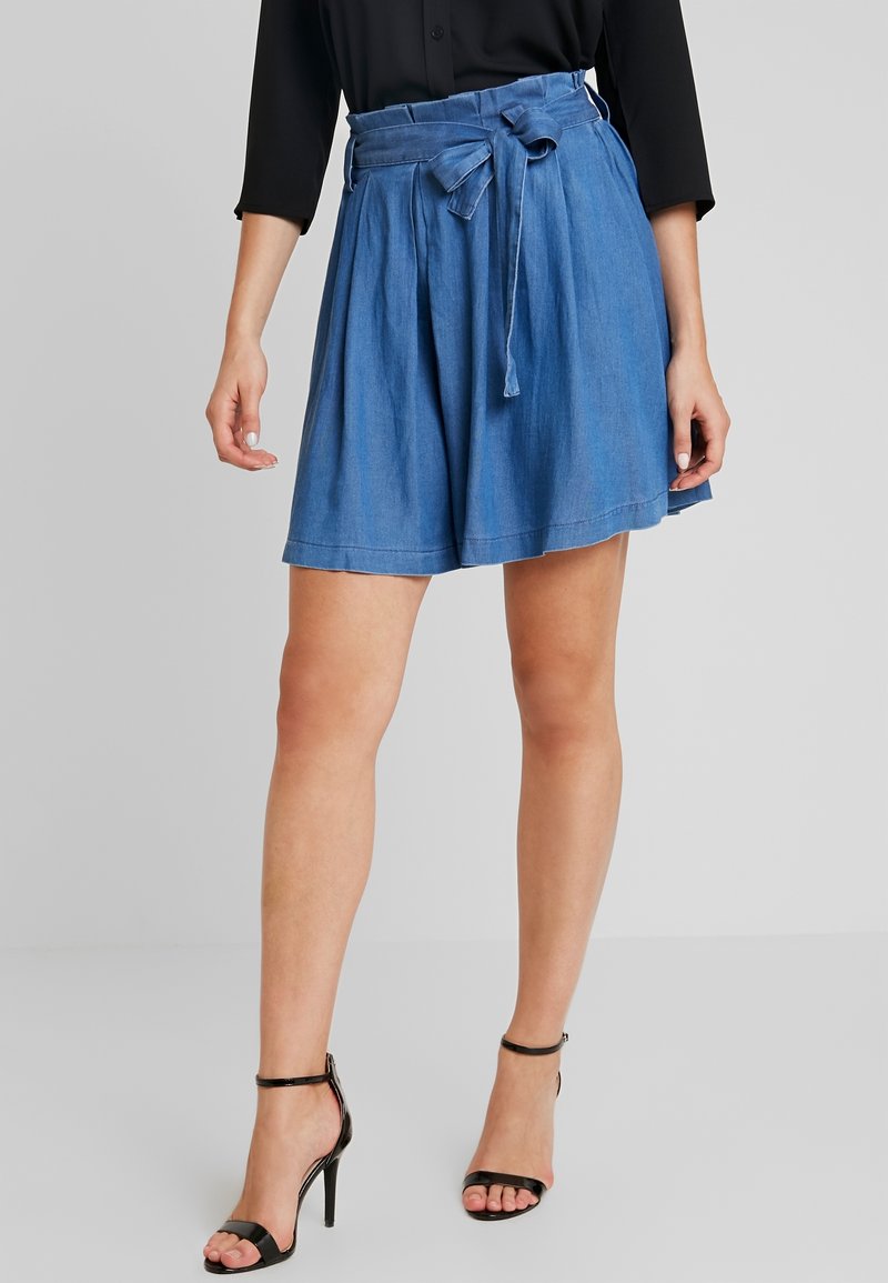 Jupe courte en denim avec des plis, taille cintrée par une ceinture, et une texture lisse. Couleur bleu clair, associée à des sandales à talons noires.