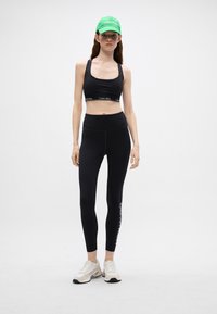 Melna sporta krūšturis ar Calvin Klein logo joslu, apvienots ar melnām augsto jostasvietu legingu, kuriem ir logo apdruka. Nēsāts ar baltām sporta kurpēm un zaļu cepuri.