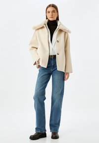 Beige ullblandad kappa med en fuskpäls-kropp, knapplåsning, kombinerad med svart polotröja, vit skjorta, blå raka jeans och svarta stövlar.