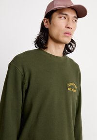 Olīvu zaļš ribots garām piedurknēm krekls ar apaļu kakla izgriezumu un dzeltenu tekstu. Valkāts ar gaismas rozā cepuri ar plakanu maliņu.