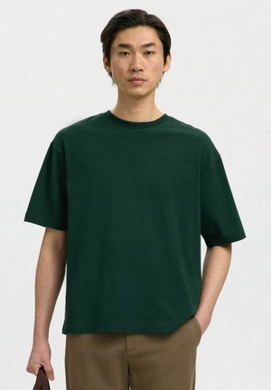 Homme portant un t-shirt vert foncé surdimensionné et un pantalon marron, debout avec une main dans la poche et l'autre tenant un objet marron.