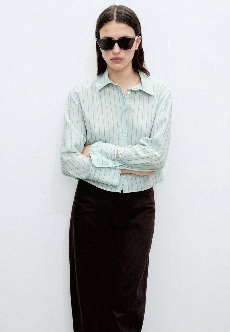 Camicia button-up a righe sottili di colore verde chiaro con colletto. Indossata con pantaloni marrone scuro a vestibilità dritta. Il modello indossa occhiali da sole neri.