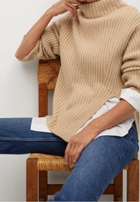 Personne assise sur une chaise en bois portant un pull côtelé beige sur une chemise blanche et un jean bleu, avec un bras levé près du visage.
