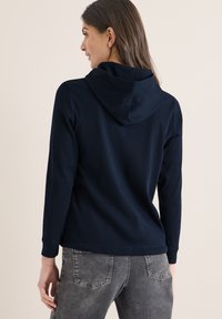 Femme portant un sweat à capuche bleu marine et un jean gris délavé, vue de dos sur un fond clair uni.