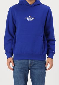 Muž v modré mikině Hilfiger s předním kapsou a logem, v kombinaci s modrými džínami, stojící před jednobarevným pozadím.