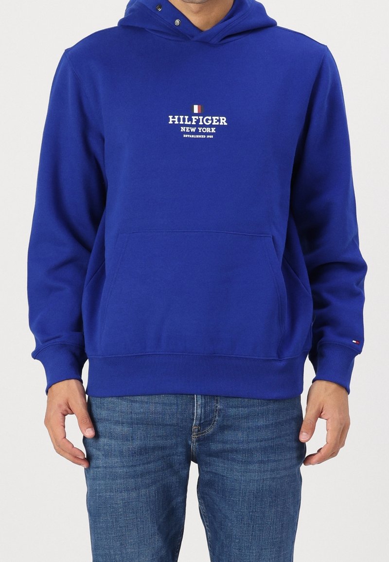 Muž v modré mikině Hilfiger s předním kapsou a logem, v kombinaci s modrými džínami, stojící před jednobarevným pozadím.