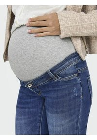 Jeans de maternidad con un acabado en denim azul, que cuentan con un cierre de botón y detalles desgastados, combinados con una camiseta gris elástica sobre una barriga redondeada.