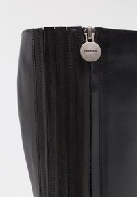 Stivaletto in pelle nera con finitura liscia, caratterizzato da cuciture verticali e una cerniera laterale con un tirante metallico circolare etichettato "GEOX."
