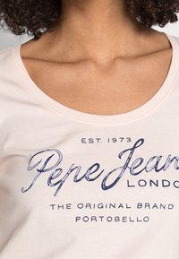 Světle růžové bavlněné tričko s kulatým výstřihem, na kterém je modrý potisk textu: "Pepe Jeans London" a další značkové detaily.