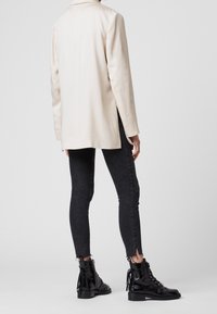 Světlý béžový dlouhý blejzr s hladkou texturou, spárovaný s úzkými černými džíny s roztrhanými lemy a černými šněrovacími kotníkovými botami s akcenty.