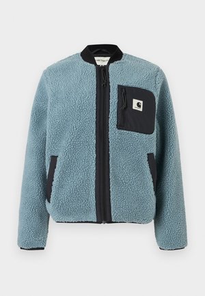 Mėlyna fliso striukė su tekstūruota paviršiumi, juodomis užtrauktukų kišenėmis, stovinčiu apykakle ir priekiniais užtrauktukais. Ant krūtinės kišenės yra logotipo pleistras.