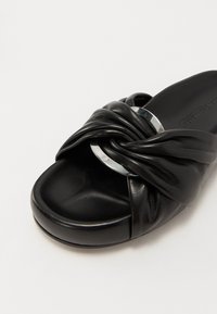 Sandale à glissière en cuir noir avec un design noué, mettant en avant un accent métallique et une texture lisse sur la partie supérieure ainsi qu'une semelle profilée.