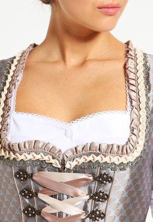 Gros plan sur une femme portant un corsage traditionnel de dirndl, orné de volants en satin et de motifs floraux, avec un chemisier blanc bordé de dentelle.