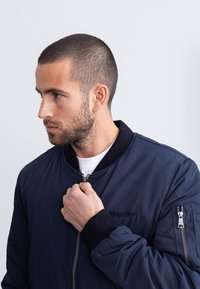 Marineblauw bomberjack met geribbelde manchetten en kraag, ritssluiting aan de voorkant en zakdetails. Gladde, duurzame stof; bevat een subtiel logo.