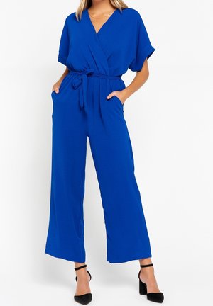 Kvinde iført en koboltblå jumpsuit med vidde ben, korte ærmer, et bånd belte i taljen, forlommer og sorte hæle med ankelrem.