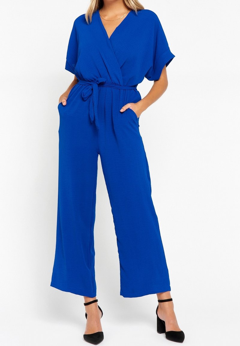 Femme portant une combinaison pantalon bleu cobalt à jambes larges, manches courtes, ceinture nouée à la taille, poches avant et talons noirs à bride cheville.