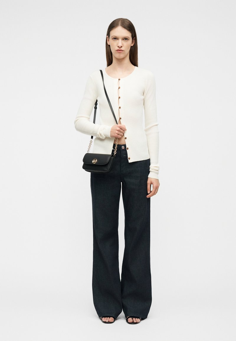 Γυναίκα που φοράει λευκό ζακέτα με κουμπιά, φαρδιά μαύρη παντελόνα, μαύρα σανδάλια, κρατώντας μία μικρή μαύρη τσάντα ώμου με χρυσή αλυσίδα.