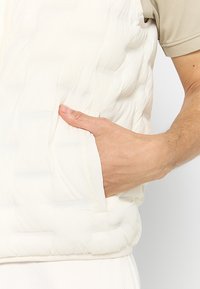 Chaleco acolchado en tejido blanco roto con textura, que incluye un bolsillo lateral y un cierre de cremallera suave.