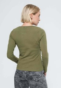 Haut à manches longues vert olive avec une texture côtelée, dotée d'un décolleté en cœur et d'un petit logo à l'arrière. Associé à un jean gris à motifs.
