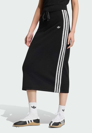 Mujer con falda midi negra de Adidas con rayas blancas a los lados, combinada con calcetines blancos de Adidas y zapatillas negro-blancas.